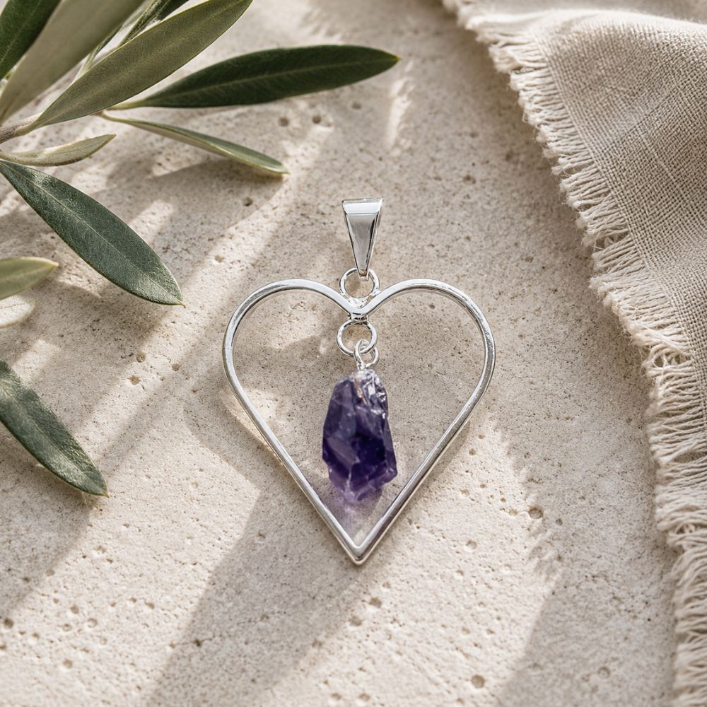 Aura Amethyst Raw Heart Pendant featuring natural raw amethyst suspended inside a silver open heart frame, displayed on a neutral stone surface with soft natural light. Each pendant is unique in shape, color, and crystal formation.