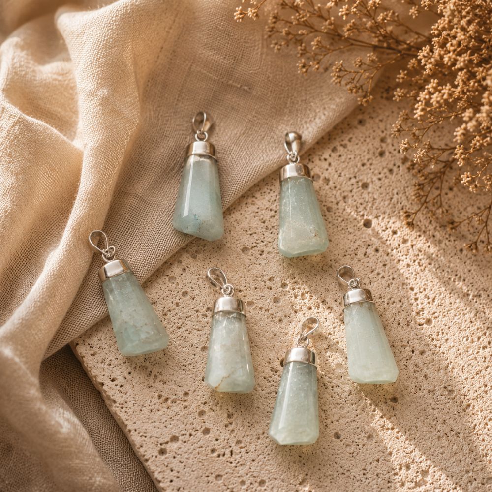 Five light green aventurine stone pendants with silver caps, arranged on a textured beige surface with draped linen and dried flowers.