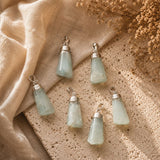 Five light green aventurine stone pendants with silver caps, arranged on a textured beige surface with draped linen and dried flowers.