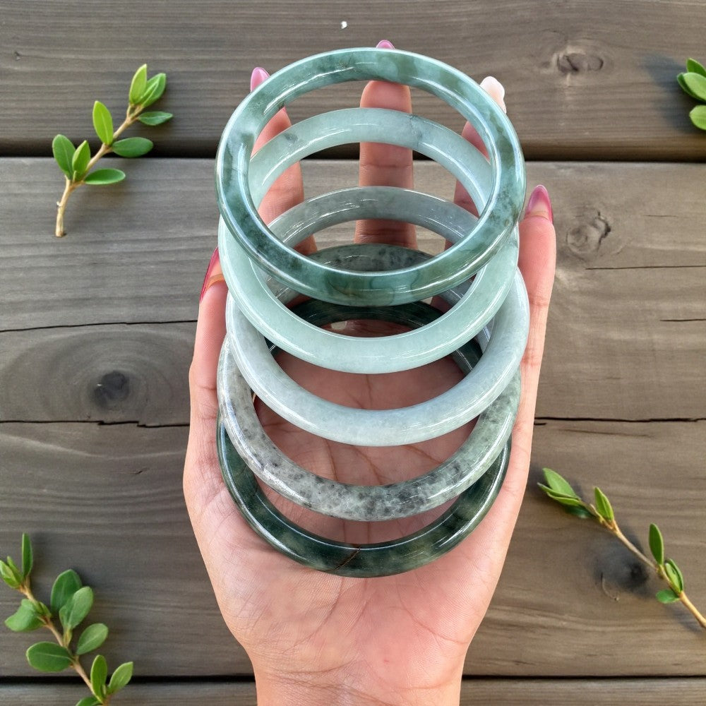 Hand holding a stack of green jade bracelets on a wooden surface with green leaves.
