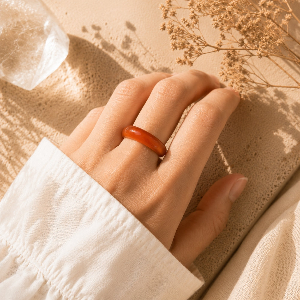A close-up of a hand wearing a smooth, rounded Carnelianring, resting on a textured beige surface with dried flowers and warm sunlight.