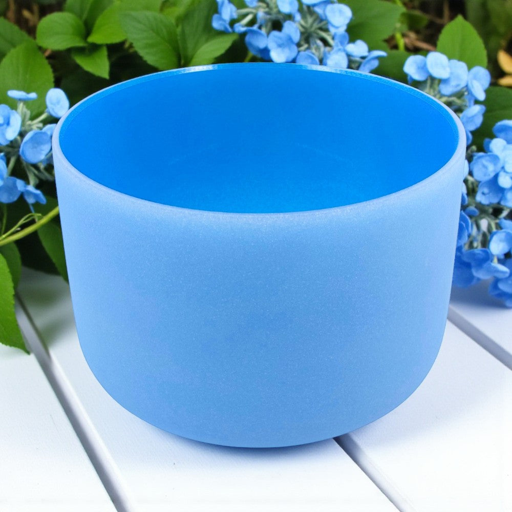 Chakra Crystal Singing Bowl - Throat on a white surface with green leaves and blue flowers in the background