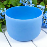 Chakra Crystal Singing Bowl - Throat on a white surface with green leaves and blue flowers in the background