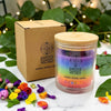 Earth's Elements Wellness candle with rainbow design and wooden lid, surrounded by flowers and leaves.