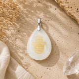 A teardrop-shaped white pendant with a gold Hamsa hand design, displayed on a textured beige surface with dried flowers and a crystal.