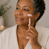 Woman using a pink facial roller on her face with a candle and plant in the background