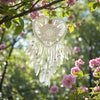 White heart-shaped dreamcatcher hanging among pink flowers and green leaves