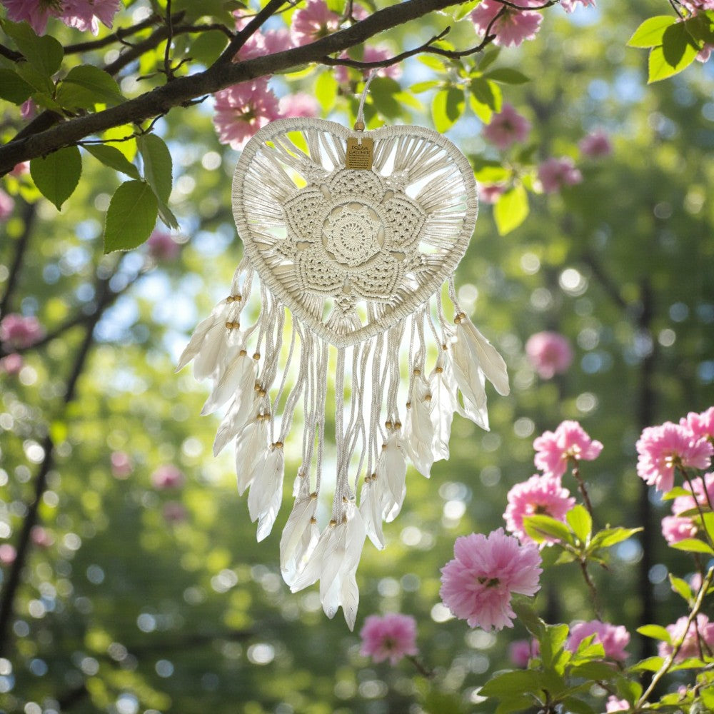 White heart-shaped dreamcatcher hanging among pink flowers and green leaves