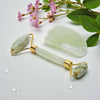 Jade facial roller and gua sha tool on a light surface with flowers and leaves.