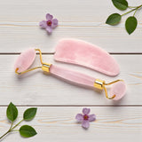 Rose Quartz facial roller and gua sha tool on a light wooden surface with flowers and leaves.