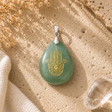 A green teardrop pendant with a gold Hamsa hand design and eye motif, resting on a textured beige stone surface with dried flowers and a clear crystal nearby.