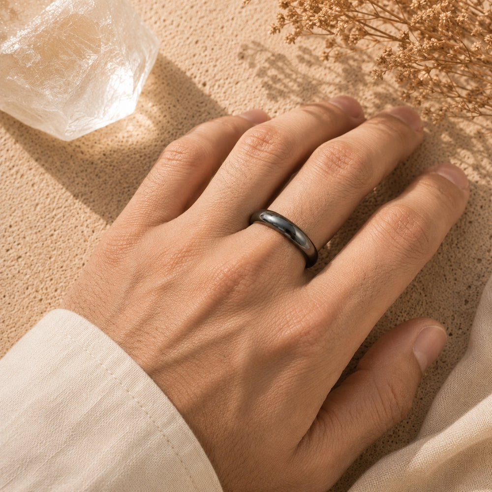 A close-up of a person's hand wearing a Hematite ring, resting on a textured beige surface with dried flowers and a clear crystal nearby.