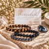 Three beaded protection bracelets displayed on a stone surface with a card reading 'Protection' and 'Earth Elements' branding, surrounded by dried flowers, greenery, and crystals. The bracelets are Tiger's Eye, Black Obsidian, and Hematite.