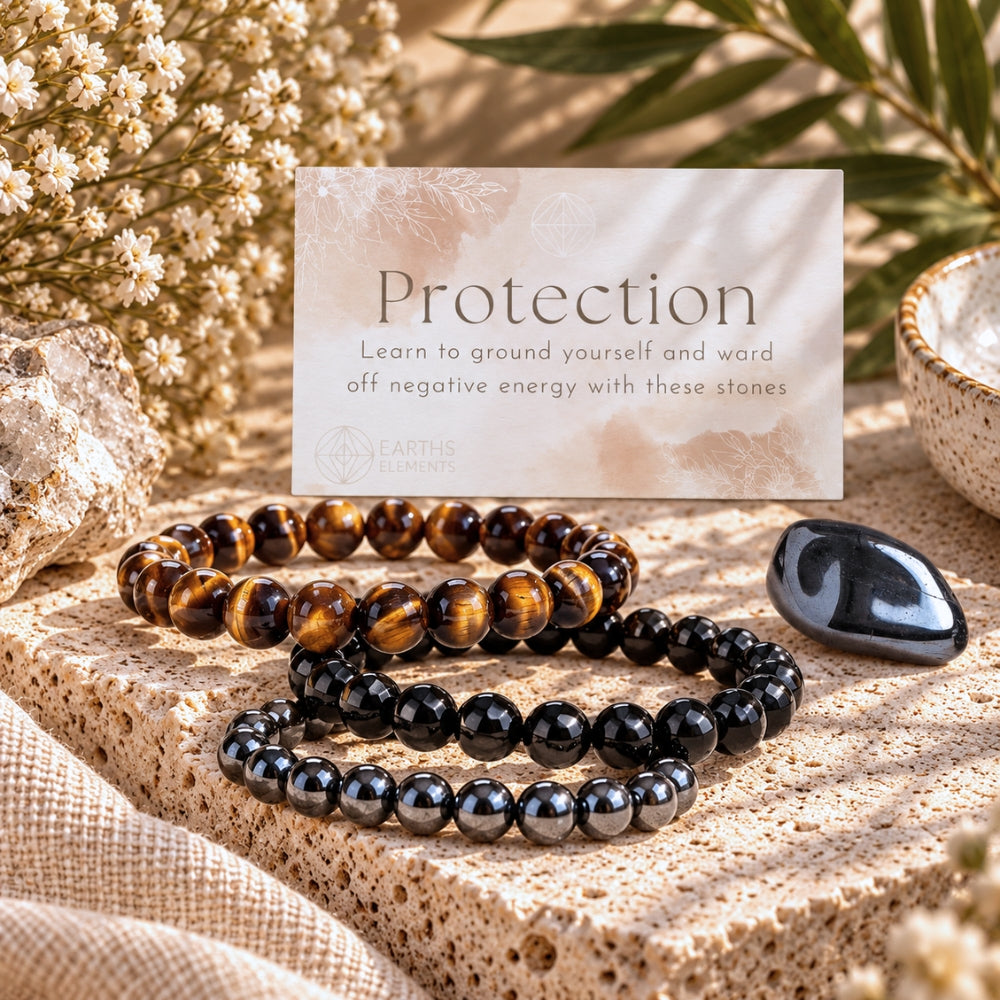 Three beaded protection bracelets displayed on a stone surface with a card reading 'Protection' and 'Earth Elements' branding, surrounded by dried flowers, greenery, and crystals. The bracelets are Tiger's Eye, Black Obsidian, and Hematite.