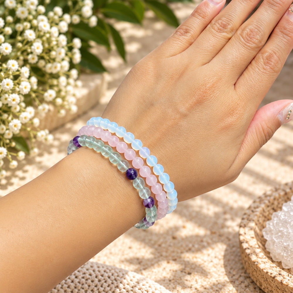 A close-up of a person's wrist wearing three beaded stretch bracelets in fluorite, rose quart, and opalite. The background is a soft-focus outdoor setting with white flowers and a woven mat.