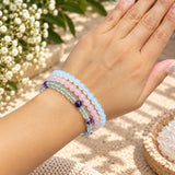 A close-up of a person's wrist wearing three beaded stretch bracelets in fluorite, rose quart, and opalite. The background is a soft-focus outdoor setting with white flowers and a woven mat.