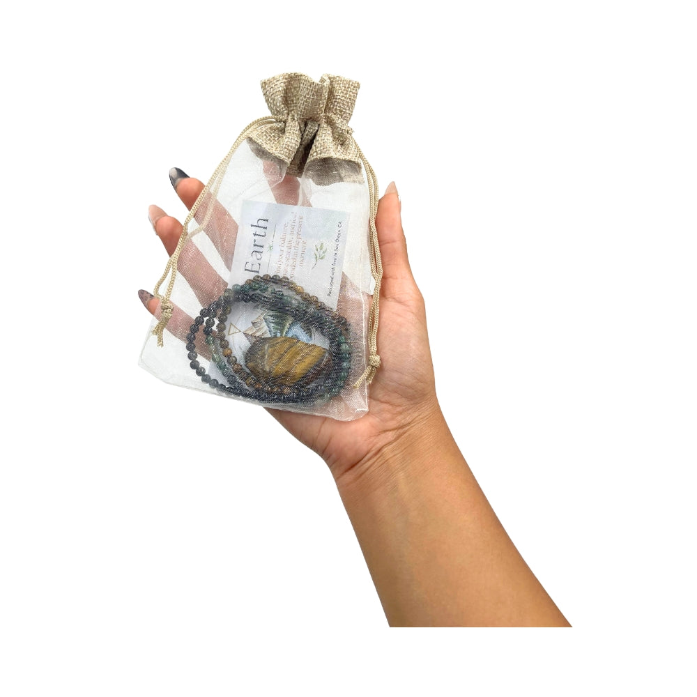 A hand holding a clear mesh drawstring pouch with a burlap-style top, containing a beaded necklace and a small card, against a plain white background.