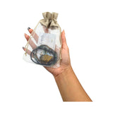 A hand holding a clear mesh drawstring pouch with a burlap-style top, containing a beaded necklace and a small card, against a plain white background.