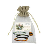 A gift set of four beaded bracelets and a polished stone displayed on a sheer white drawstring pouch with a printed label reading 'Earth'. The bracelets are made of moss agate, black obsidian, and tiger's eye with a tiger's eye tumble.