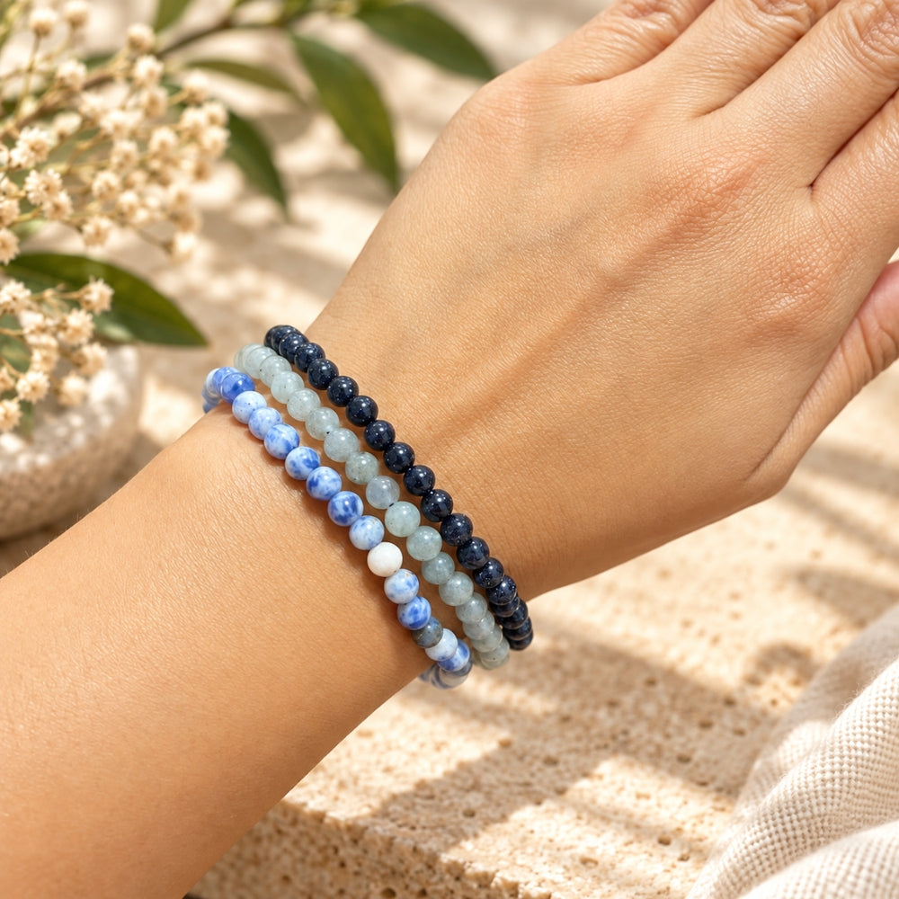 Three beaded stretch bracelets in shades of navy, light blue, and marbled blue-and-white, worn on a person's wrist against a warm beige textured background with soft floral bokeh.