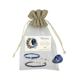 Moon phase crystal gift set in a sheer white drawstring pouch with a beige fabric top, featuring two beaded bracelets, a polished blue moon stone, and a printed card.