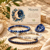 Three beaded stretch bracelets in shades of navy, light blue-gray, and deep navy are arranged on a sandy surface beside a blue stone and a printed card featuring a moon illustration and the word 'Moon'.