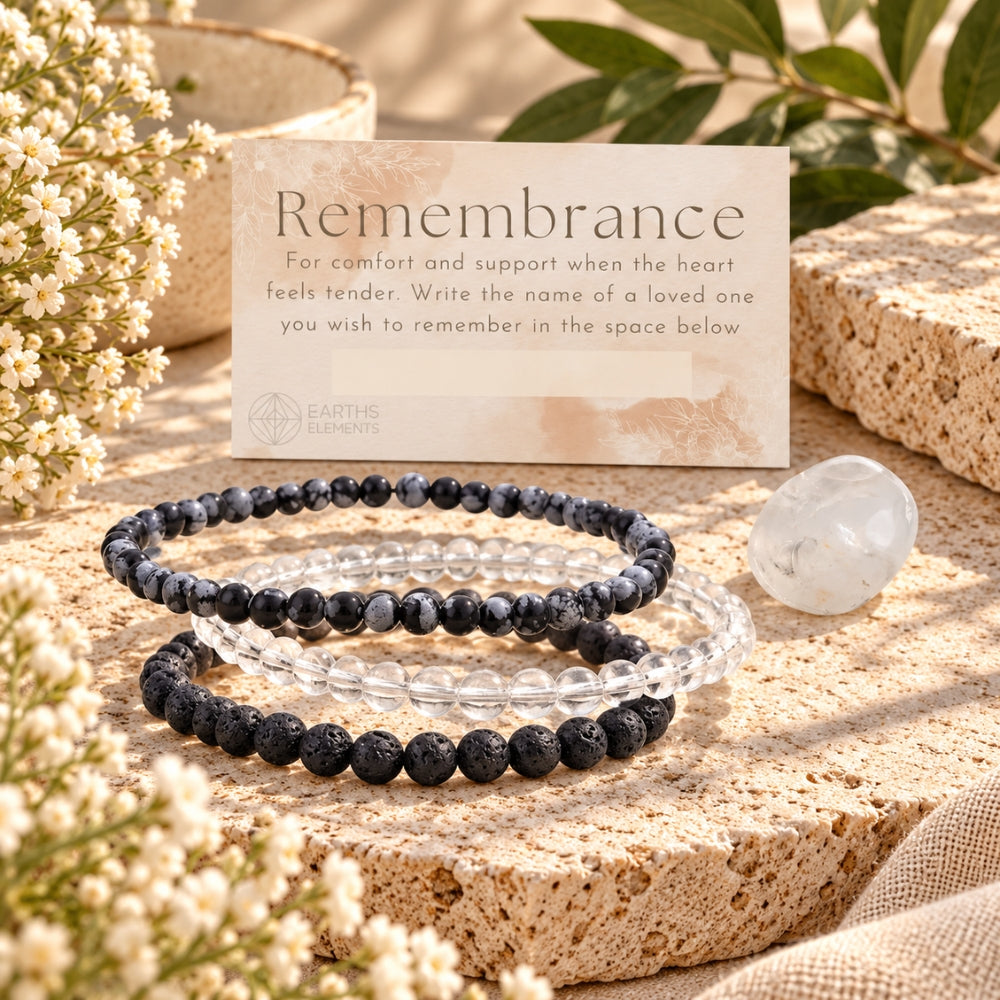 Three beaded stretch bracelets displayed on a textured stone slab with white flowers and green leaves. The bracelets are black lava stone, clear quartz, and snowflake obsidian.