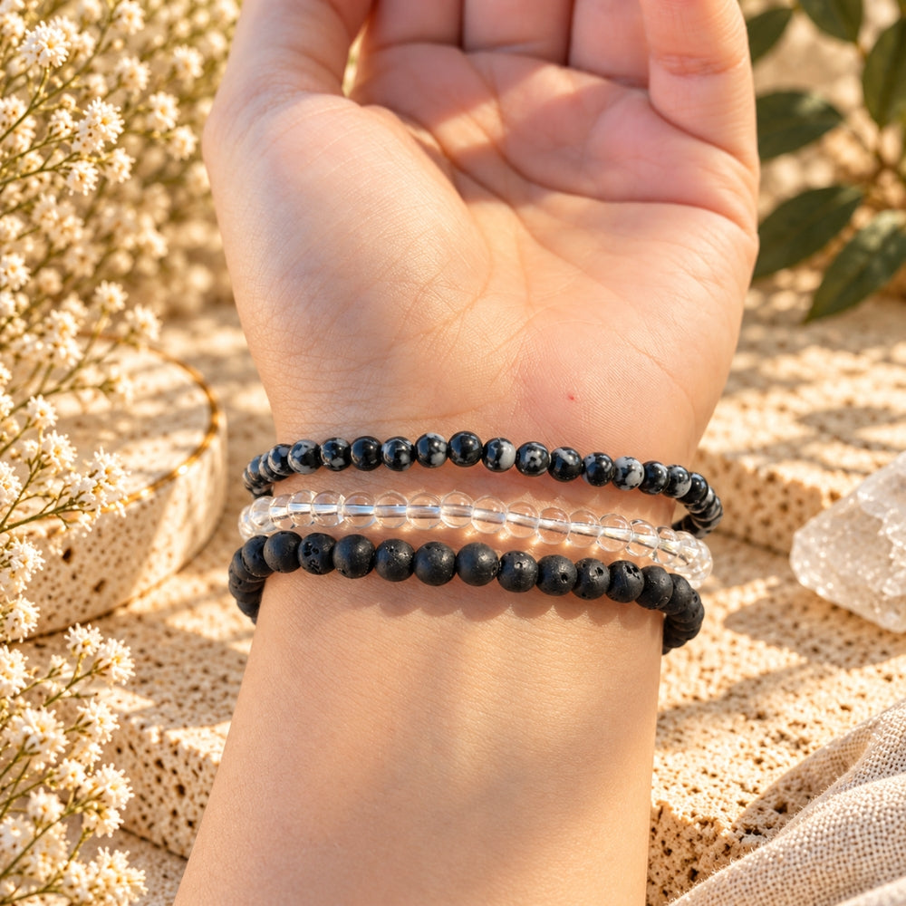 A close-up of a person's wrist wearing two beaded bracelets:lava stone, clear quartz, and snowflake obsidian, set against a warm sandy background with dried flowers.