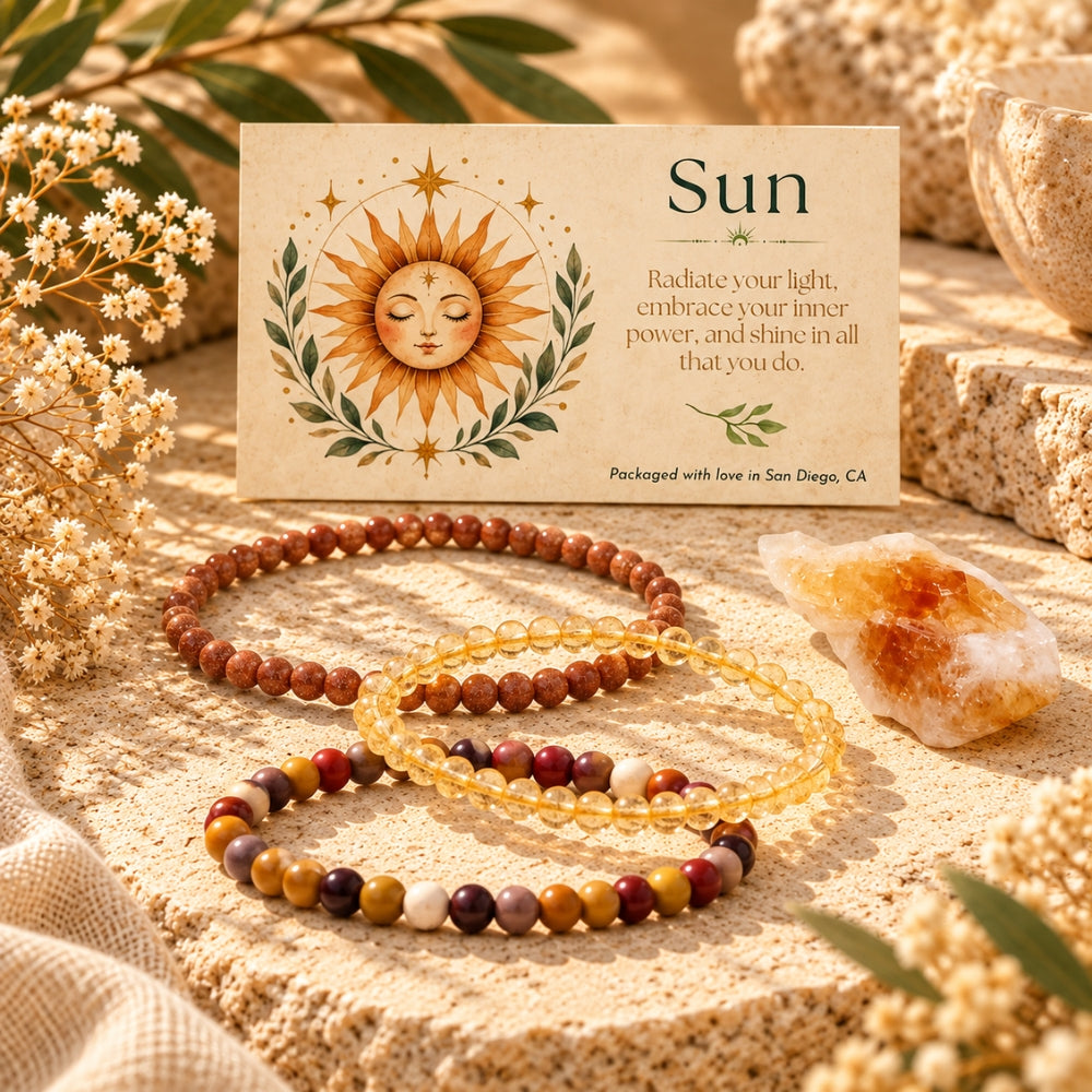 Three beaded stretch bracelets arranged on a textured stone surface beside a printed card labeled "Sun" with a sun illustration. The bracelets includeCitrine, Goldstone, Mookaite and a crystal citrine cluster around them.