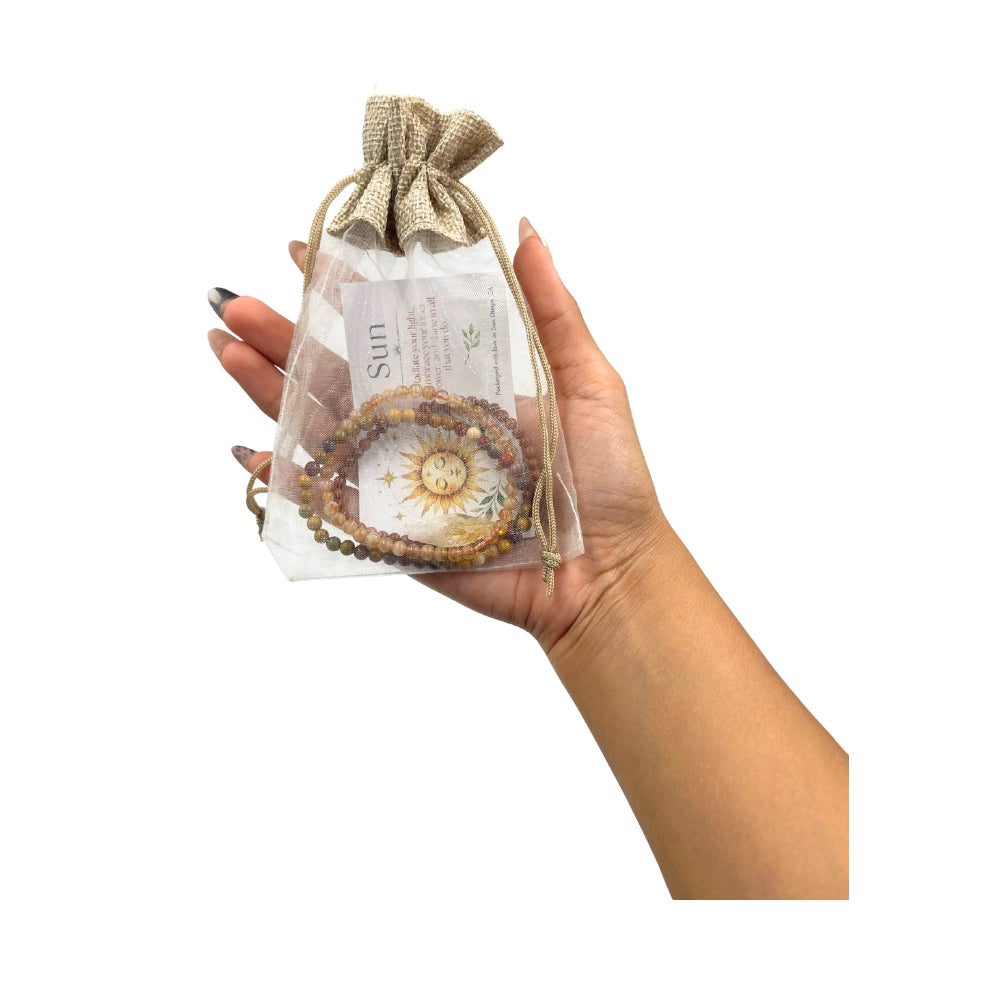 A hand holding a clear drawstring pouch with a burlap-style top, containing a beaded bracelet and a printed card labeled 'Sun'.