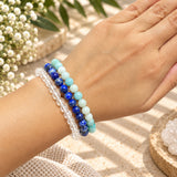 Three beaded stretch bracelets worn on a person's wrist, featuring blue lapis lazuli, amazonite, set against a styled background with dried flowers and a woven textile.