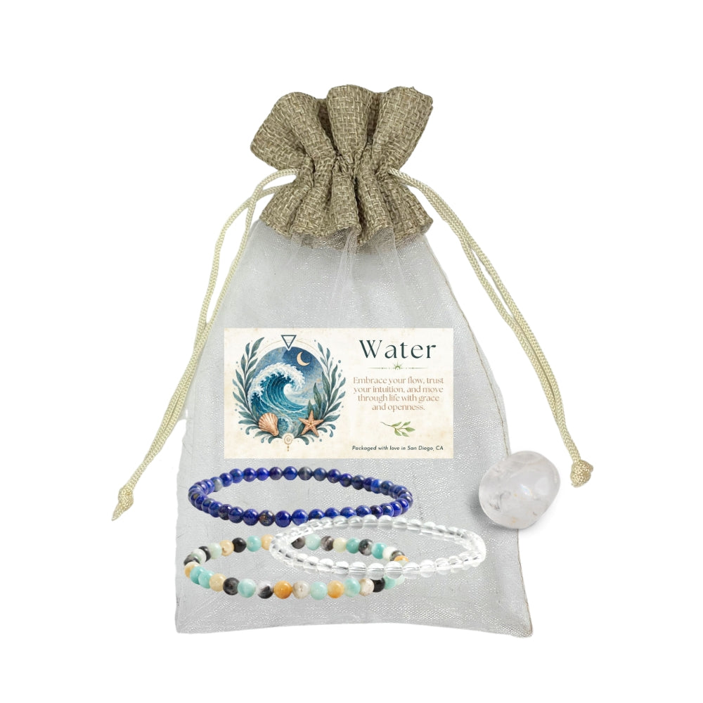 A beaded bracelet gift set labeled 'Water' displayed inside a sheer light gray drawstring pouch. The set includes a dark blue bead bracelet, a clear crystal bracelet, and a multicolor beaded bracelet, alongside a clear quartz tumble and an illustration card.
