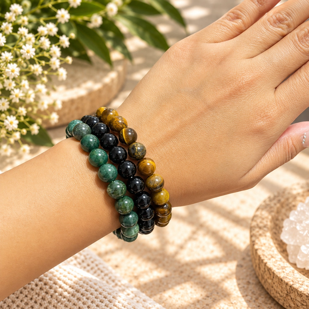 Three beaded stretch bracelets worn on a person's wrist, featuring moss agate, black obsidian, and tiger's eye, set against a warm, sunlit background with flowers and woven textures.