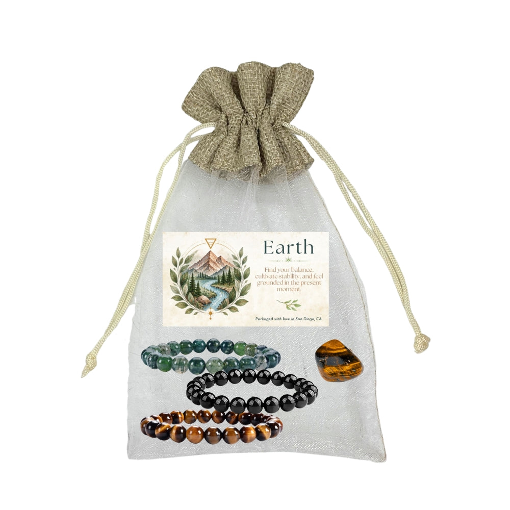 Earth Balancing crystal set in a sheer white drawstring pouch with a kraft paper top. The set includes moss agate, black obsidian, and tiger's eye, and a printed card with a mountain illustration and the text 'Earth'.