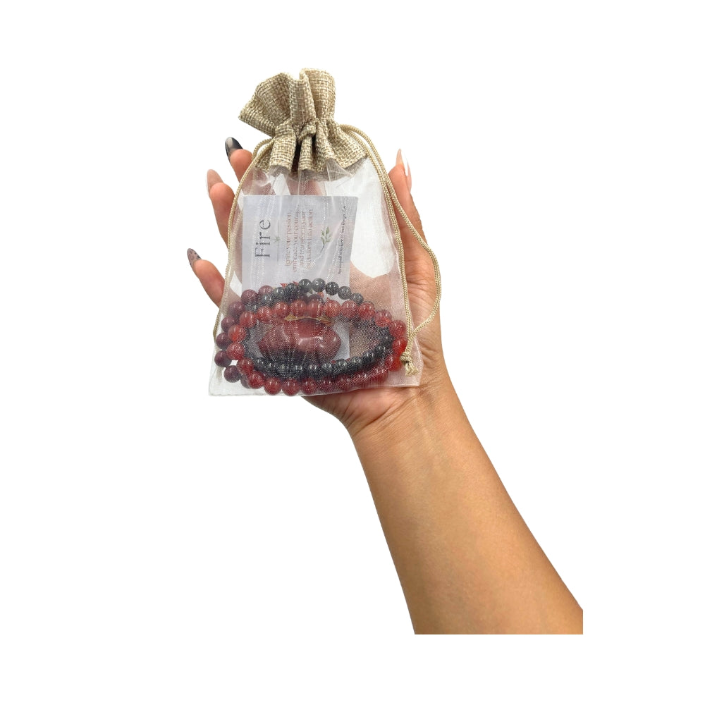 A hand holding a clear drawstring pouch with a burlap-style top, containing a red and dark gray beaded bracelet set with a central carved stone, against a plain white background.