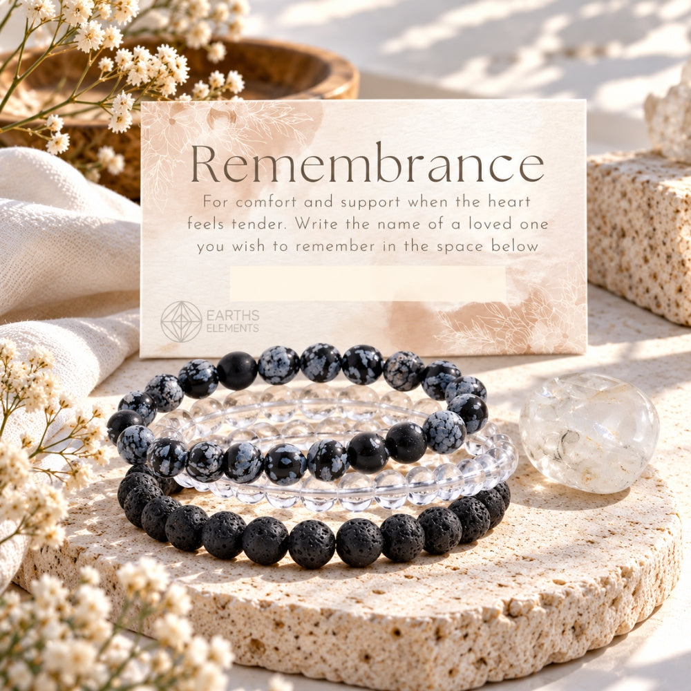Three beaded bracelets displayed on a stone slab with a card reading 'Remembrance' and the brand 'Earth's Elements.' One bracelet is made of black lava stones, and the other is made of clear quartz and snowflake obsidian.