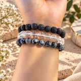 Three beaded stretch bracelets worn on a person's wrist. The top bracelet is made of dark black lava stones, the middle bracelet is made of clear quartz beads, and the bottom bracelet is made of snowflake obsidian.