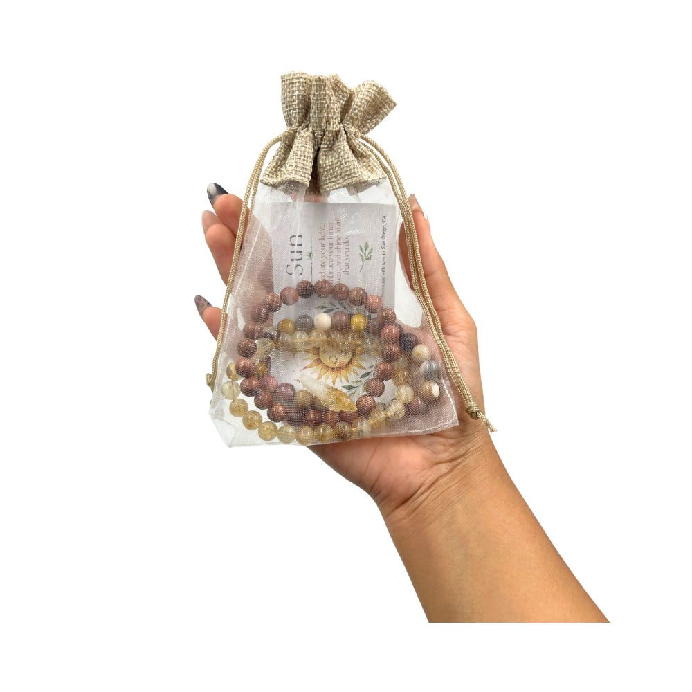 A hand holding a clear drawstring pouch with a burlap-style top, containing goldstone, citrine, and mookaite beaded bracelet or set of bracelets and a printed card, against a plain white background.