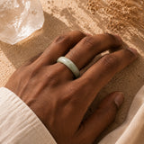 A light green jade bangle ring worn on a person's hand, resting on a textured sandy surface with dried flowers and a crystal cluster nearby.