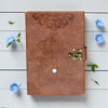 Brown leather notebook with embossed design and gold latch on a light wooden surface with blue flowers.