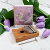A kalimba thumb piano with a wooden body, metal tines, and a metal wrench, styled on a white marble surface with purple tulips and green leaves, with its illustrated pink and purple box behind it.