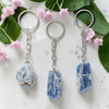 Three keychains with blue stone pendants on a marble surface with pink flowers and green leaves.