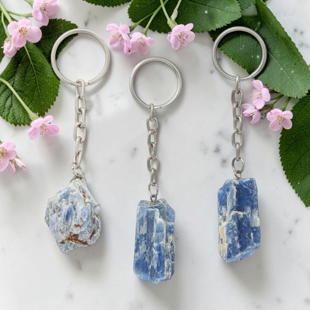 Three keychains with blue stone pendants on a marble surface with pink flowers and green leaves.