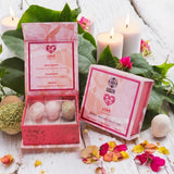Pink packaging with tumbled crystals and candles on a wooden surface with flowers and leaves.