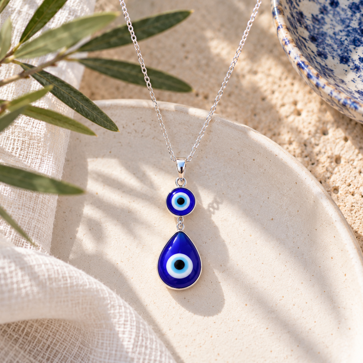 Small blue Evil Eye necklace featuring a round charm above a teardrop pendant, suspended from a delicate silver chain on a cream ceramic surface.