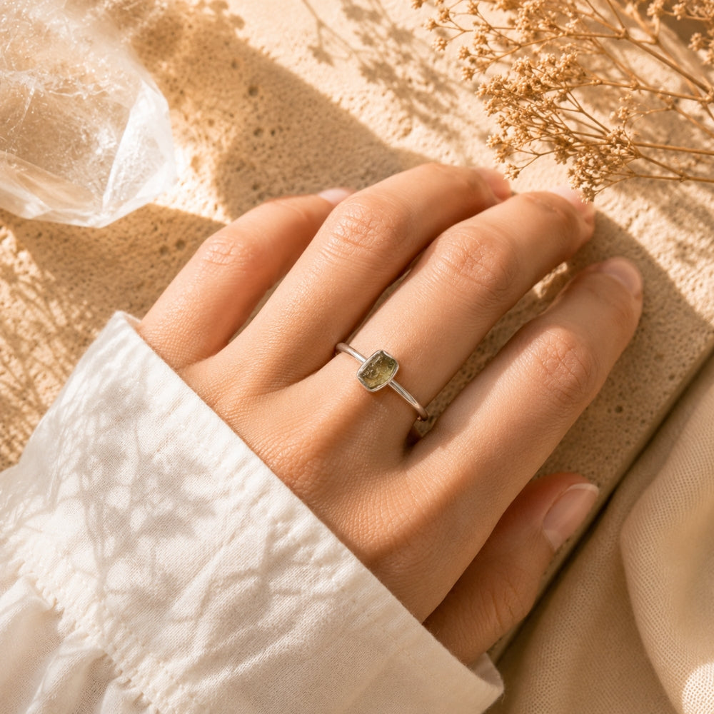 A close-up of a hand wearing a delicate silver ring with a small rectangular Moldavite, resting on a textured beige surface with dried flowers and soft shadows.