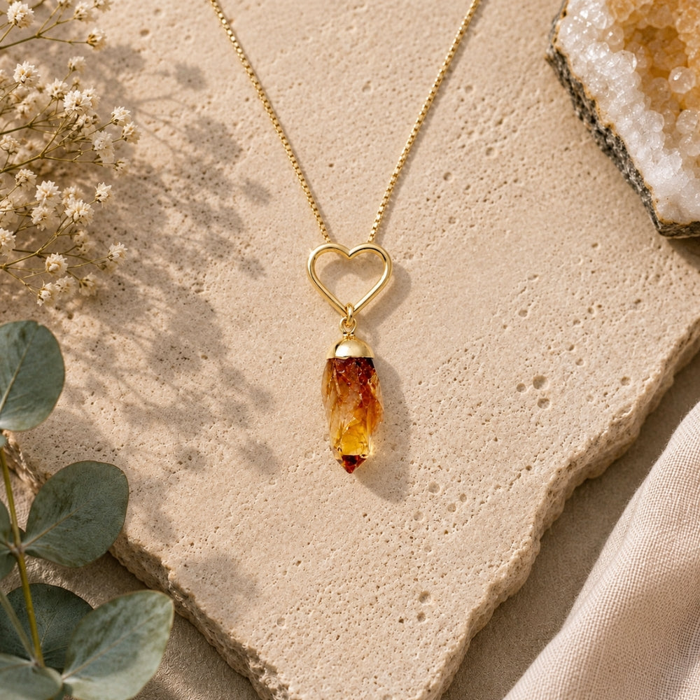 A gold heart-shaped pendant necklace with a raw citrine crystal drop, styled on a textured beige stone slab with eucalyptus leaves and dried flowers.