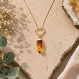 A gold heart-shaped pendant necklace with a raw citrine crystal drop, styled on a textured beige stone slab with eucalyptus leaves and dried flowers.