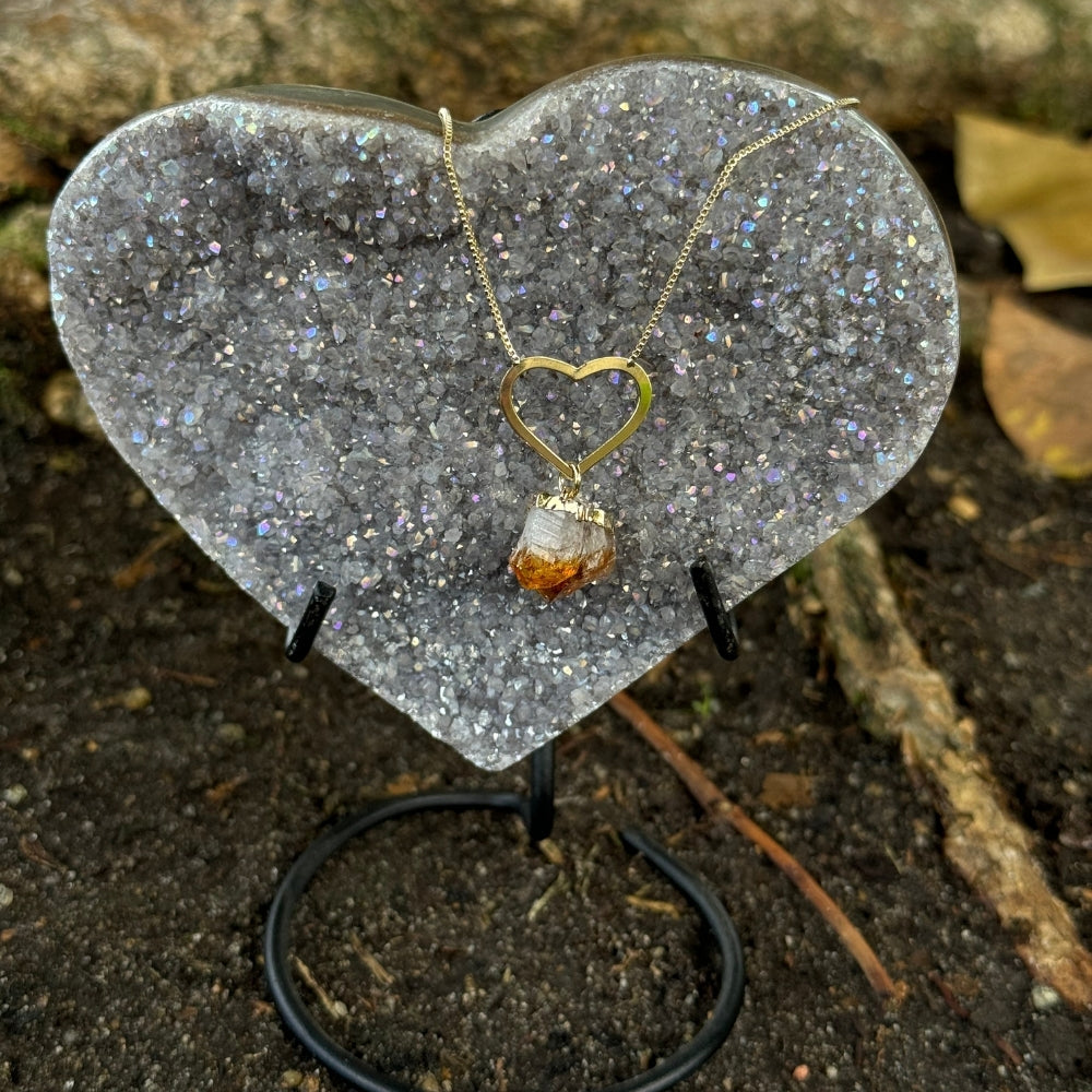 A gold heart-shaped pendant necklace with a raw amber crystal hangs from a chain on a heart-shaped glitter display stand, set outdoors on dark soil with twigs and leaves in the background.