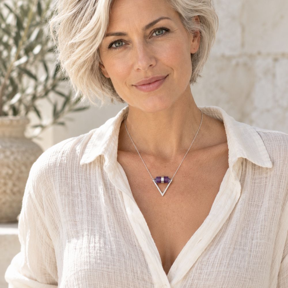 Silver-haired woman wearing an Amethyst Double Terminated Triangle Necklace with a purple crystal and silver geometric pendant, styled with an ivory linen blouse in soft natural daylight.